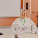Photograph of an elderly person wearing a white sweatshirt and a green lanyard with an ID badge, seated indoors against a peach-colored wall.