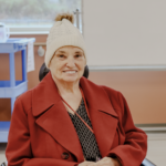 Photograph of an elderly person seated indoors wearing a red coat, patterned shirt, and white knit hat with a pom-pom.